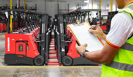 A worker in a warehouse full of forklifts holds a clipboard and pen