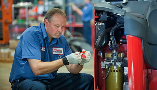 A field service technician works on a Raymond forklift, inspecting a part in his hands.