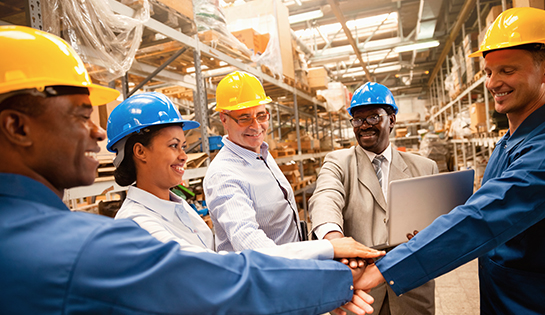 Five people wearing hardhats in a warehouse each with a hand in the middle of their group, symbolizing team work