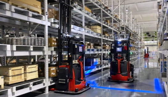 Two AGVs in a warehouse aisle. One is picking a pallet, the other is moving down the aisle with the blue safety light aimed at the ground