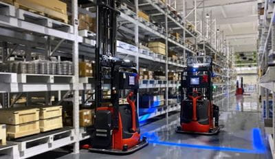 Two AGVs in a warehouse aisle. One is picking a pallet, the other is moving down the aisle with the blue safety light aimed at the ground